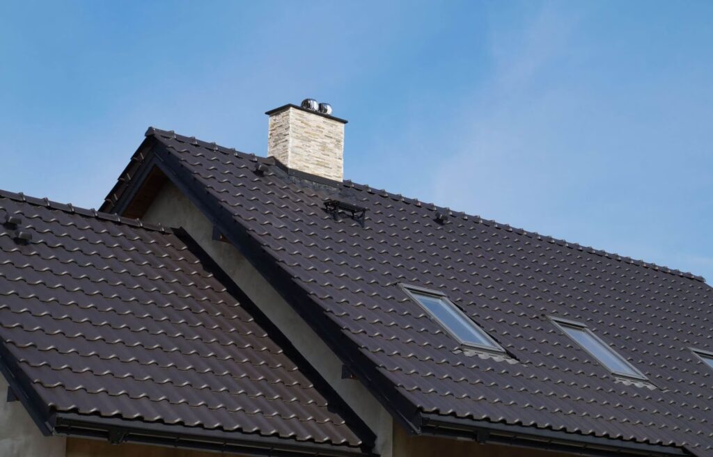Toit en tuiles foncées d’une maison, avec cheminée et fenêtres de toit, sous un ciel bleu.