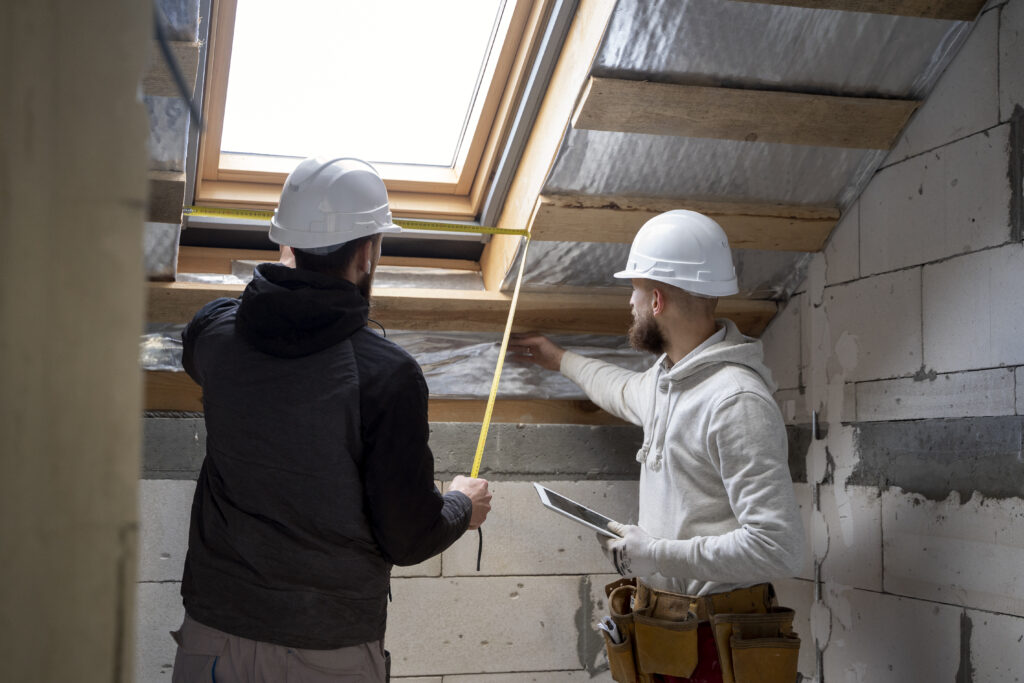Deux artisans en casques de chantier mesurent une zone sous toiture près d’une fenêtre de toit avec un mètre ruban, l’un tenant une tablette, dans un grenier en cours d’aménagement.